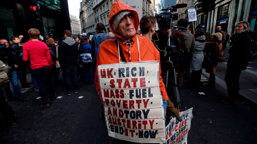 Man stands amid protestors wearing a sign: "UK rich state, mass, fuel, poverty, and Tory, austerity, end it now.