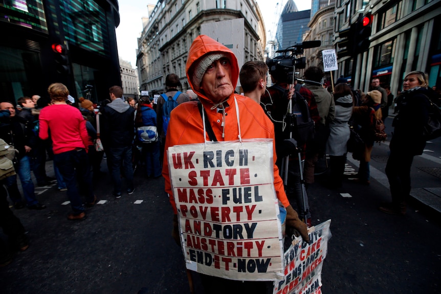 Man stands amid protestors wearing a sign: "UK rich state, mass, fuel, poverty, and Tory, austerity, end it now.