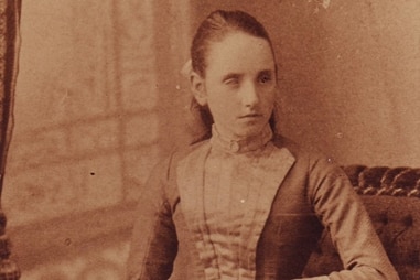 Photo of Tilly Aston as a young girl of 16 sitting in a chair with a handbag in her lap. 
