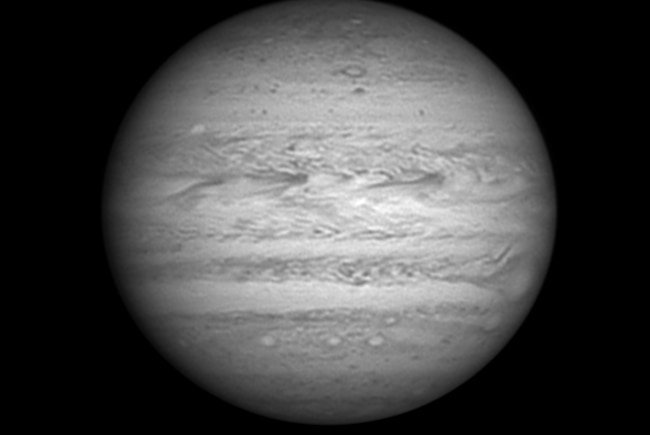 A close up image of Jupiter in black and white showing a small white spot in the top left side.