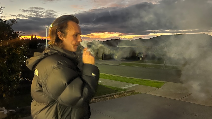 A man in a puffer jacket in a suburban street exhaling after using an e-cigarette