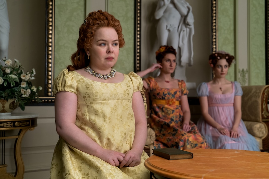 A curvy woman wearing a yellow period gown, with two women in the background