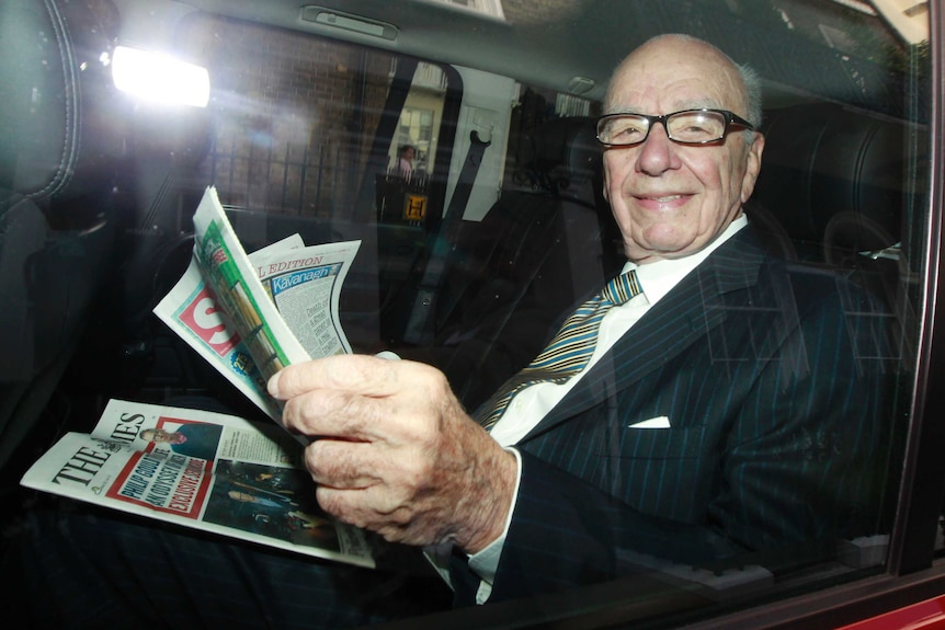 Rupert Murdoch holds copies of The Sun and The Times newspapers as he looks towards camera in the back of a car