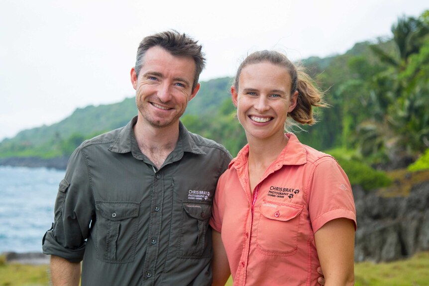 A medium shot of Chris in a grey shirt and Jess in a pink shirt on a tropical coastline smiling at the camera.