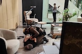 An armed man kneels on the floor next to a person who is lying on the floor with their hands behind their back, inside a shop