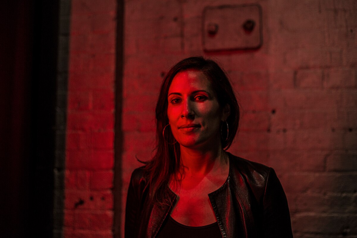 The director stands against a bare cement wall, lit in dramatic red and shadows.