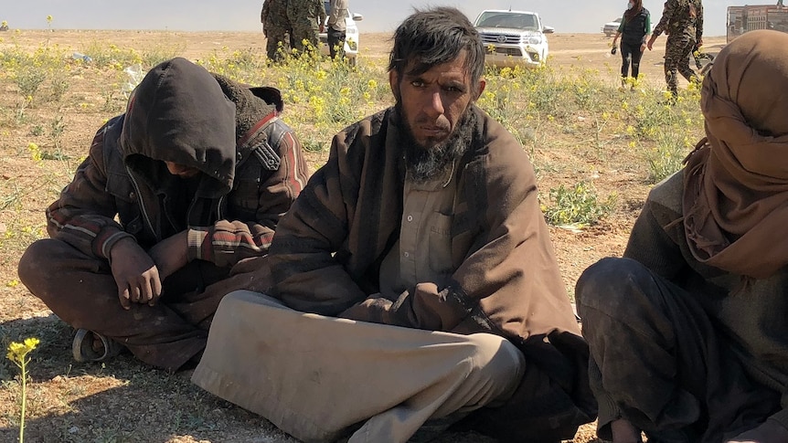 Three men sit on the ground while a group of people in military fatigues talk in the background.