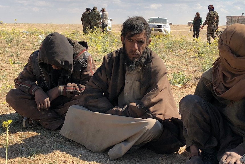 Three men sit on the ground while a group of people in military fatigues talk in the background.