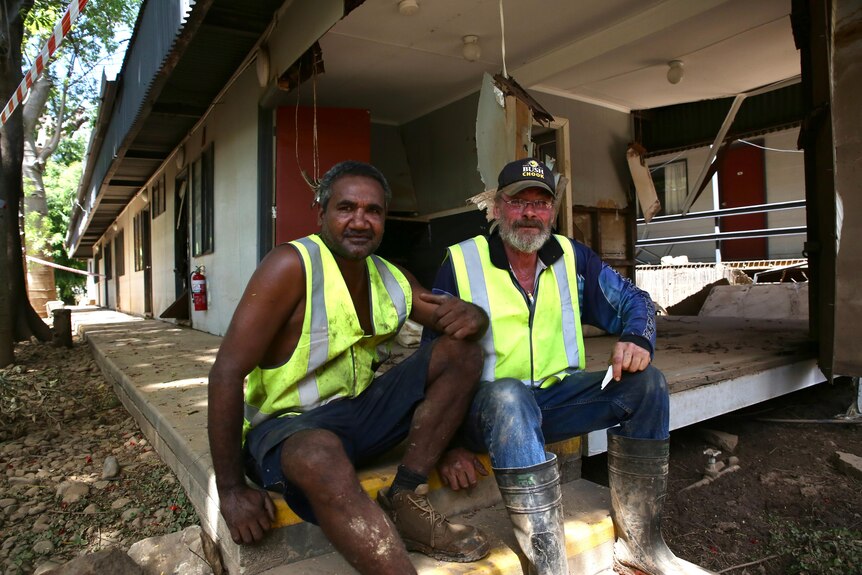 Dos hombres con equipo de alta visibilidad se sientan afuera de una propiedad dañada por la inundación. 