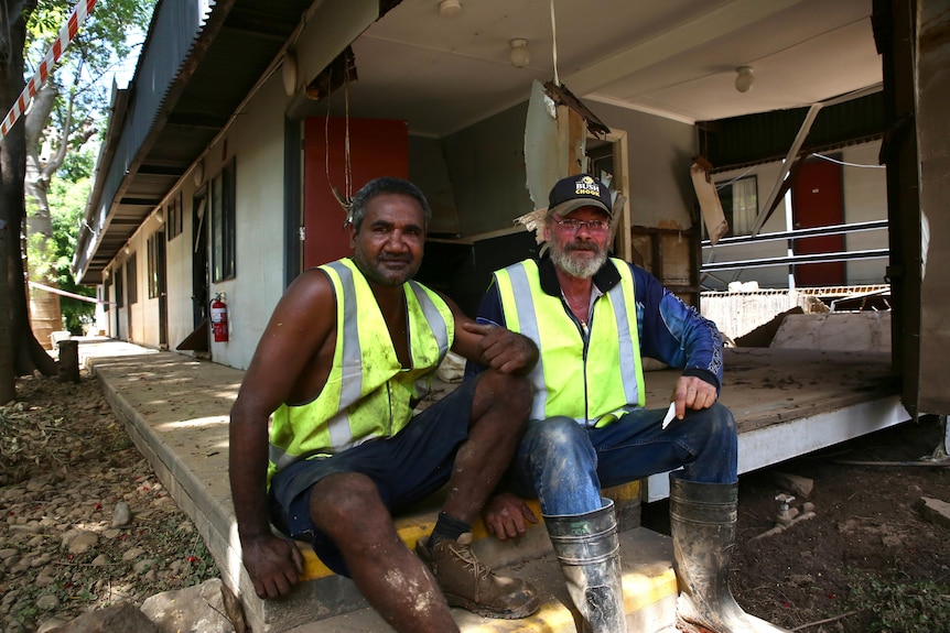 Deux hommes portant des vêtements à haute visibilité sont assis à l'extérieur d'une propriété endommagée par les inondations. 