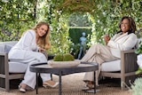 Adele and Oprah both wearing light-coloured suits, laughing together while sitting on garden furniture. 