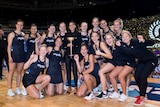 A group of netballers kneel and stand, smiling, on the court with the Constellation Cup trophy.