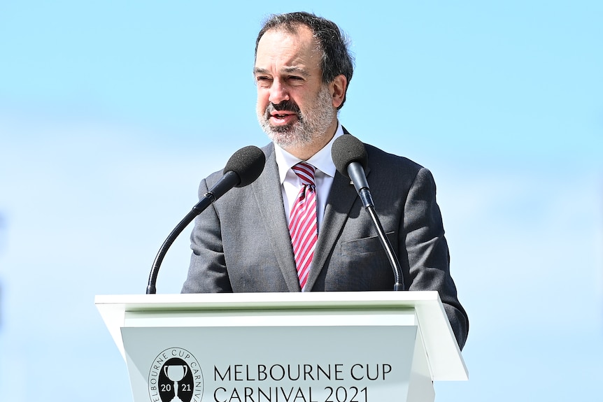 A state government minister speaks into a microphone at a lectern with the words "Melbourne Cup Carnival 2021" on it.