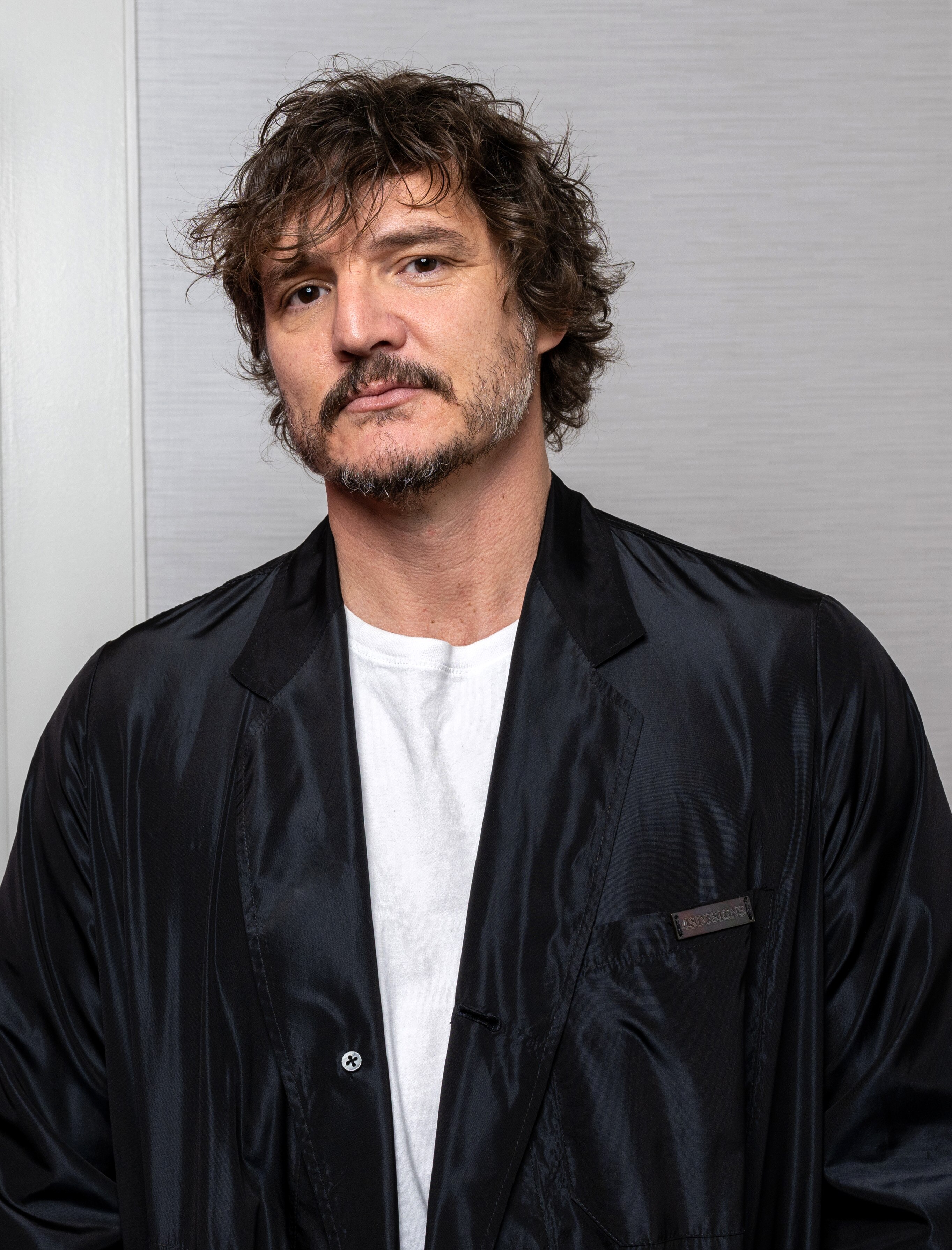 Pedro Pascall dressed casually in a black jacket with white shirt, curly brown hair, moustache, beard