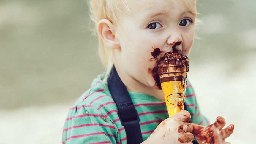 a small blond child looks at the camera, his face covered in chocolate as he takes an enormous bite out of an ice-cream cone
