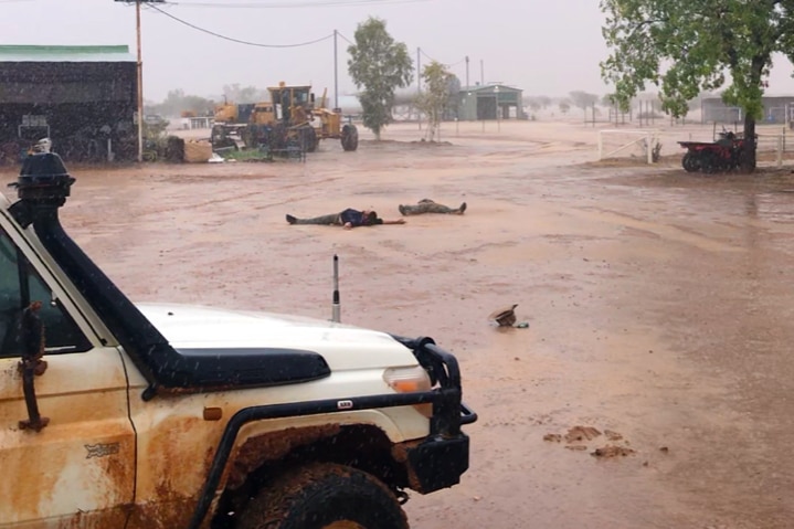 Two men make mud angels in the rain at Roxborough Downs Station with farm equipment in the background