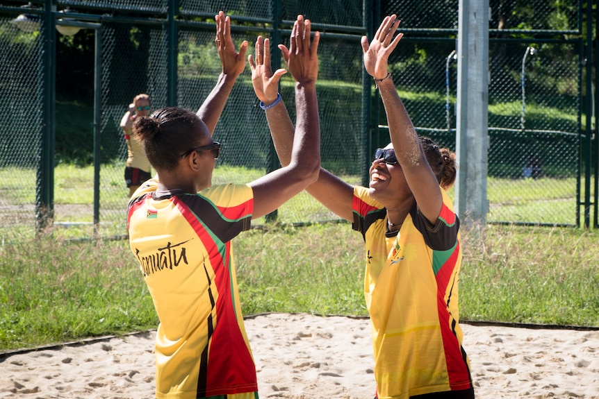 Two women on beach raise hands toward each other.