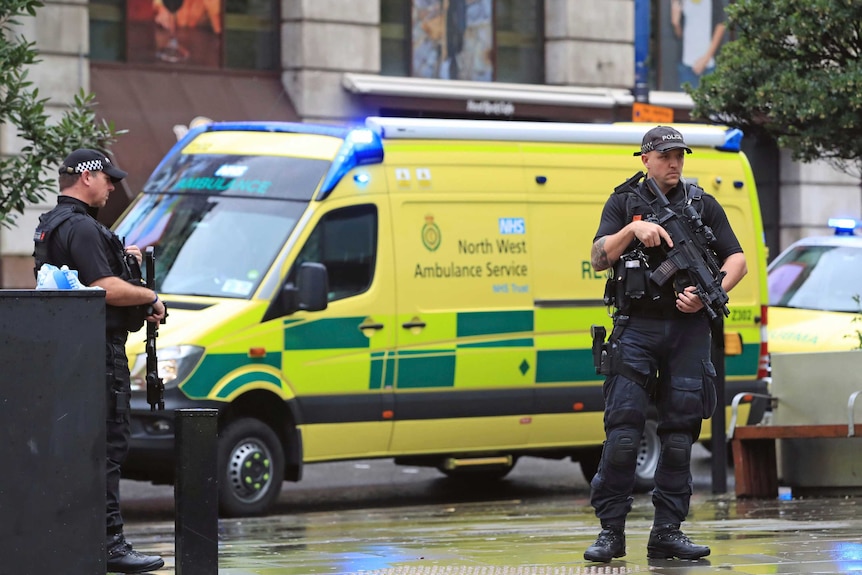 Heavily armed UK police stand in front of a yellow ambulance.