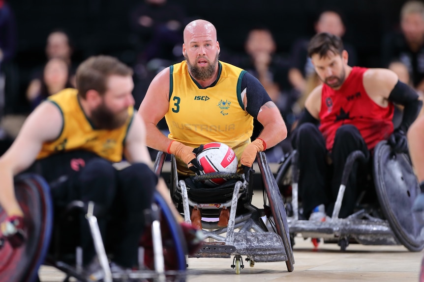 An Australian wheelchair rugby player holds the ball in his lap while he sits in his chair on the court during a game.