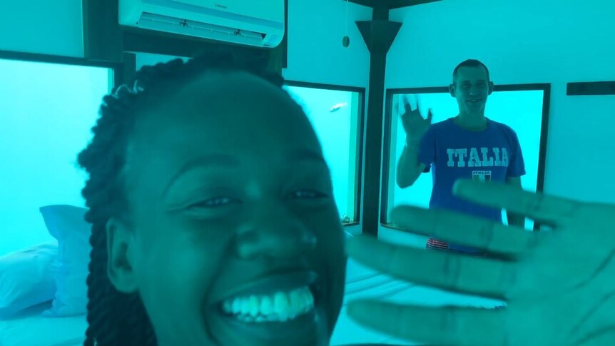 A woman, close to the camera, and a man, in the background, wave at the camera while standing in an underwater bedroom.