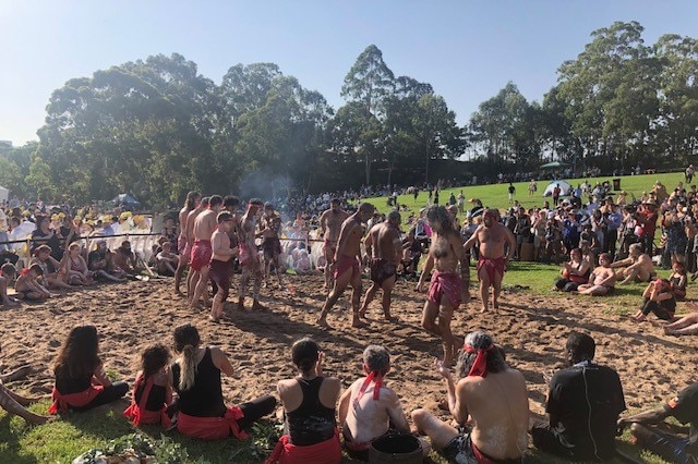 Aboriginal dancers gathered in a park - some are standing in the middle of a circle while others are sitting around the edge.