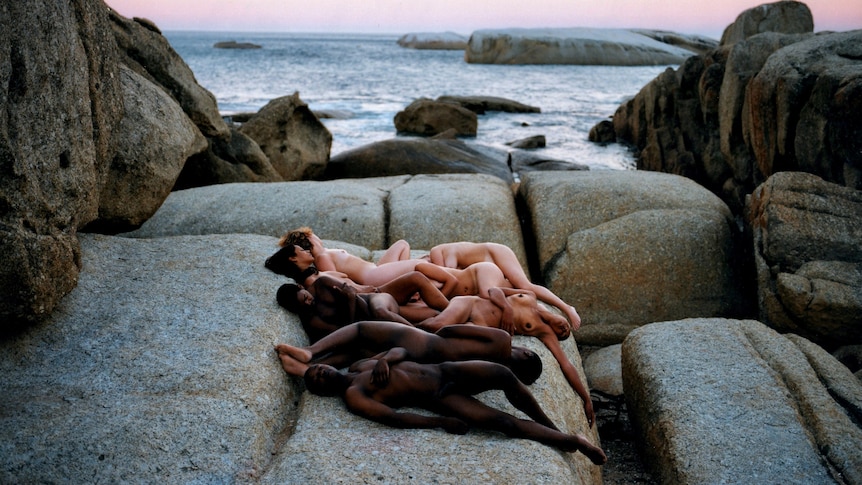 A group of naked people with black, brown and white skin arranged together on a rock with the ocean behind