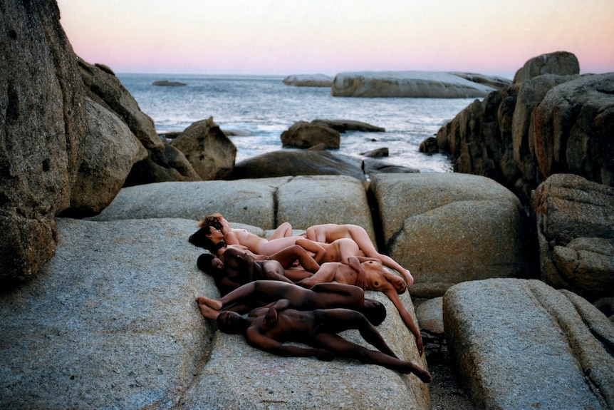 A group of naked people with black, brown and white skin arranged together on a rock with the ocean behind