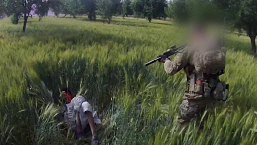 A soldier with a blurred face points a gun at a man lying prone in a field.