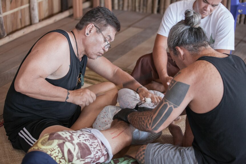 Traditional tattooists sit around man as he lays face down, being tattooed with the 'au. 