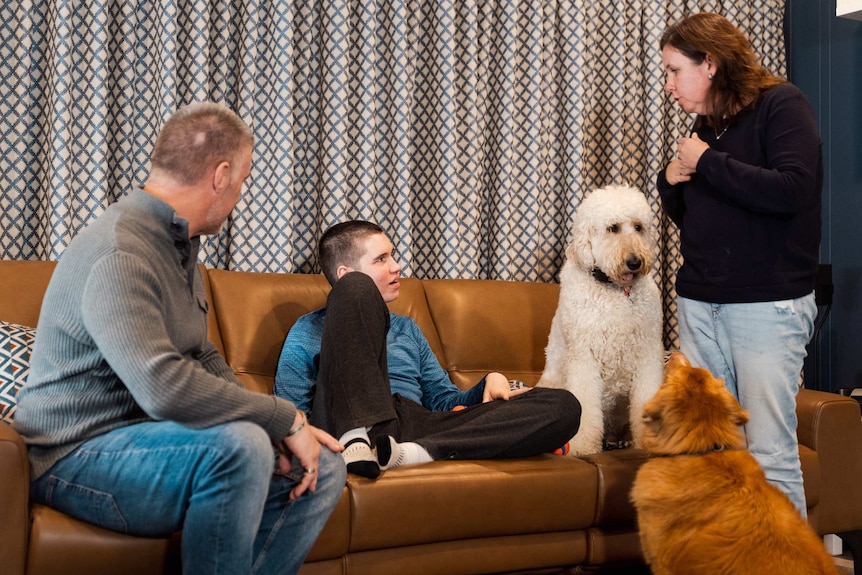 A teenage boy sits on a couch with his father to his right and his mother and two dogs to his left.