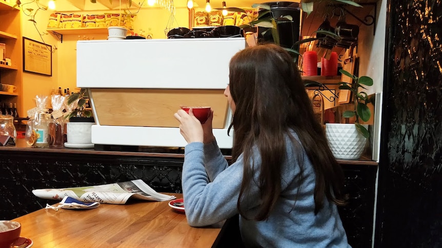 A woman is seen sitting in a cafe holding a coffee cup while looking straight ahead of herself. Her face is hidden.