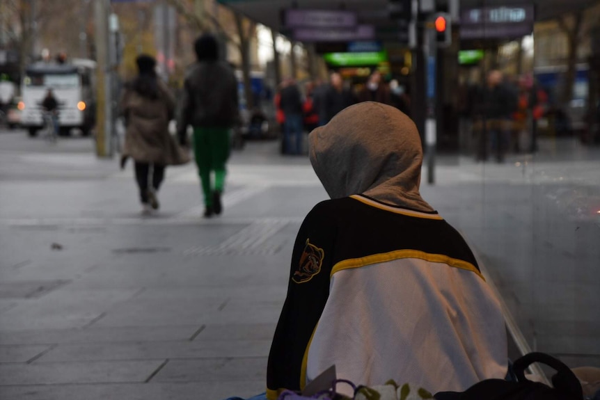 A homeless man sits on a blanket on a footpath wearing a hoodie.