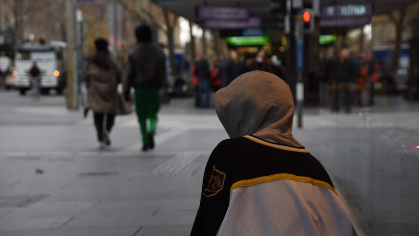 A homeless man sits on a blanket on a footpath wearing a hoodie.