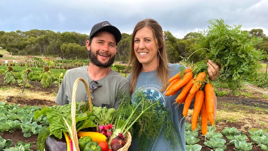 a happy couple, man and woman holding vegetables like carrots smiling in front of a garden.