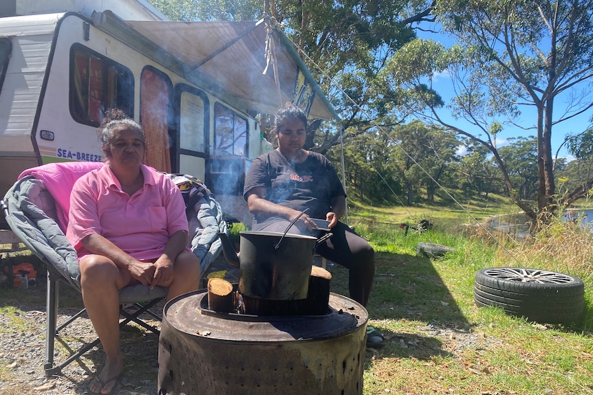 Two women sit outside an old caravan in front of an open fire.