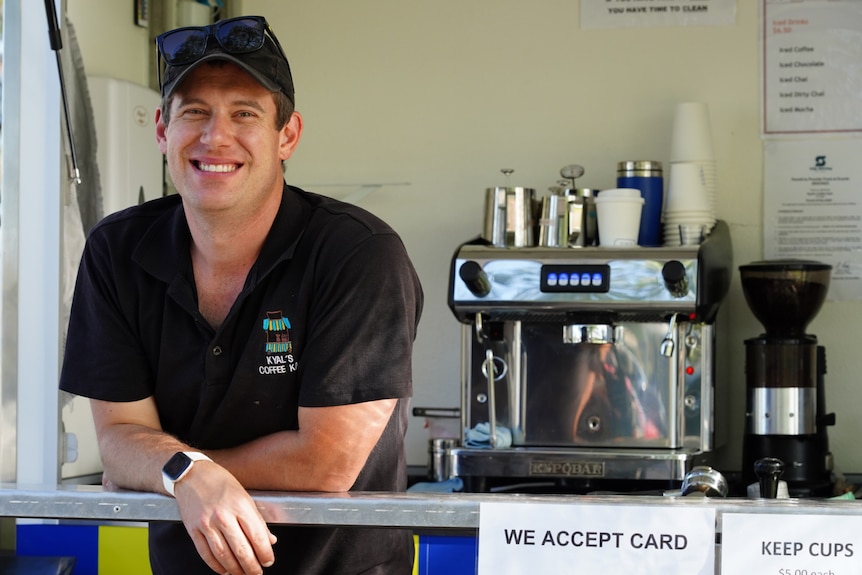 Man wearing a black shirt and cap in a cart with a coffee machine behind him.