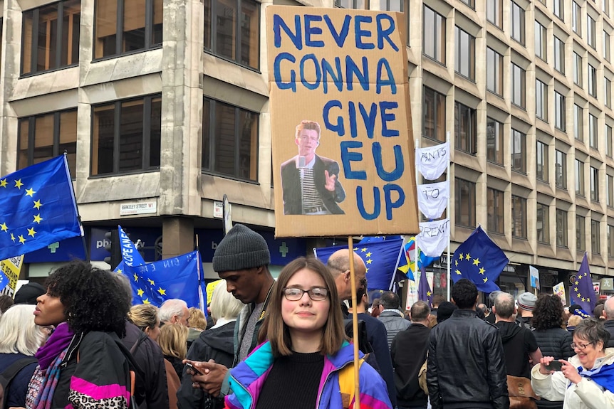 A young woman holds up a cardboard sign with a crowd of people around her