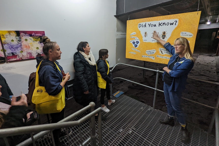 A blonde white woman pointing at an informational sign about bees and other people are looking.