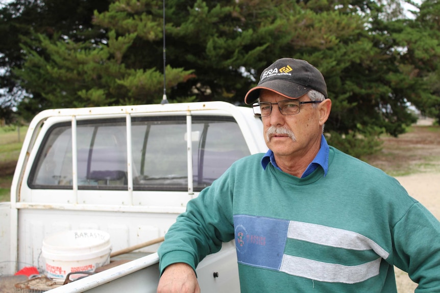 A farmer wearing glasses and a cap leans over the back of his ute with a green background
