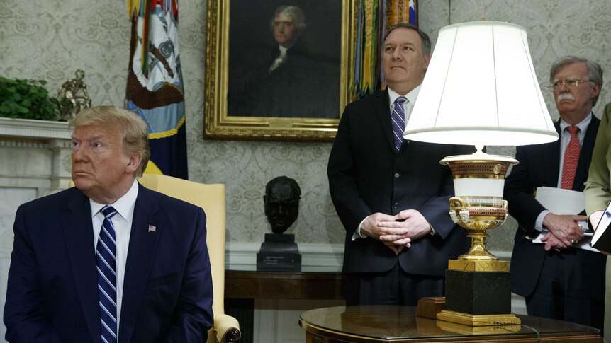 Donald Trump sits on a chair while Mike Pompeo and John Bolton stand behind him.
