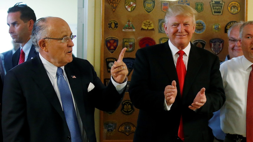 A man in a suit points his left index finger up as another man stands nearby smiling and clapping.