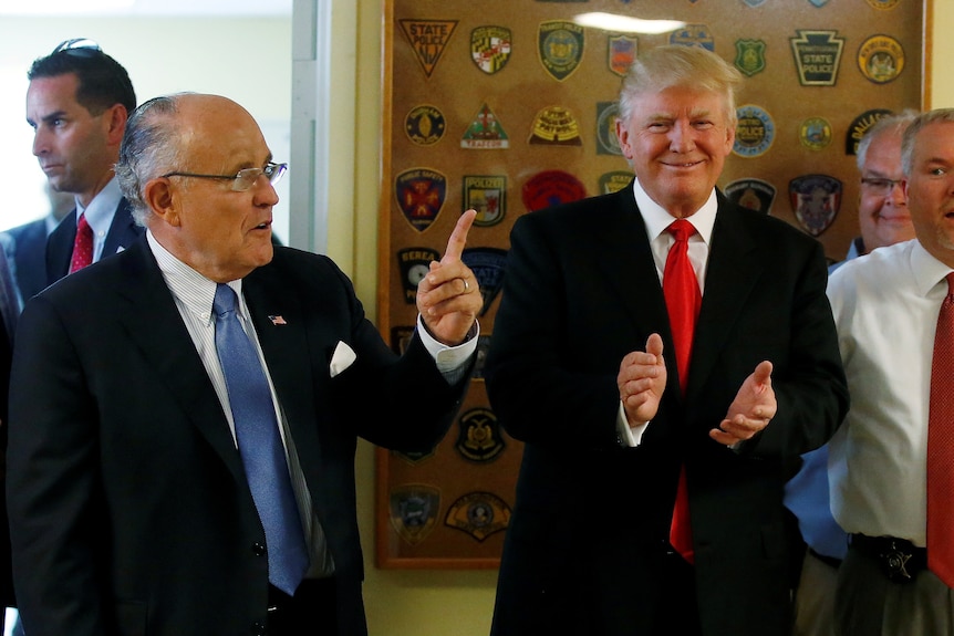 A man in a suit points his left index finger up as another man stands nearby smiling and clapping.