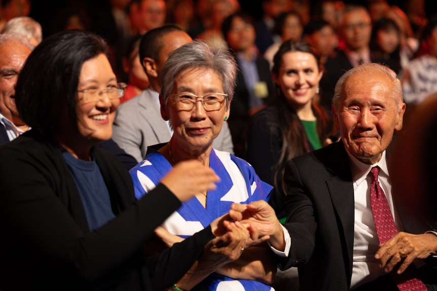 An older man is seen smiling at the camera, sitting next to two younger women.