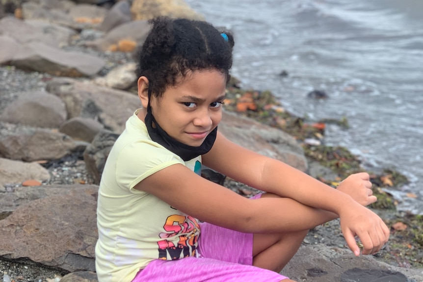 A young girl sits on rocks on shoreline in Fiji.