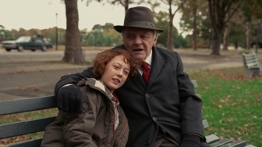 Young white boy in brown jacket sits beside elderly white man in fedora and black coat on a park bench.