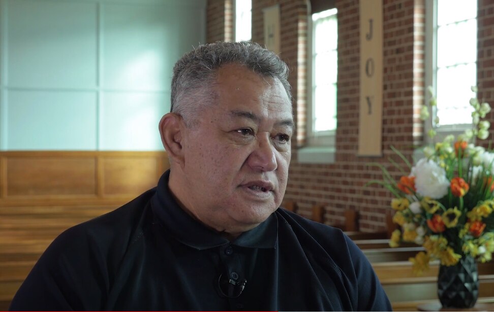 A Pacific Island man with short grey hair wearing a black button-up top sitting in a church pew