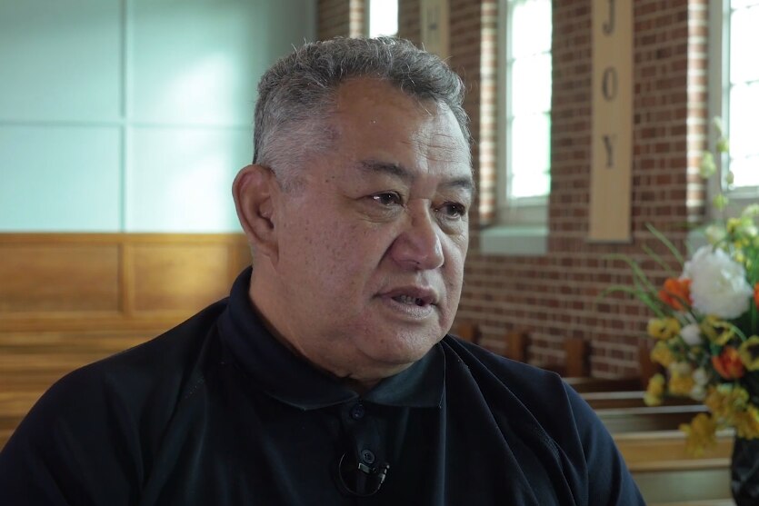 A Pacific Island man with short grey hair wearing a black button-up top sitting in a church pew