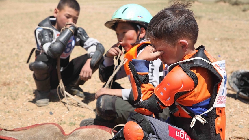 Three child jockeys sits on the ground. One holds his forehead and closes his eyes.