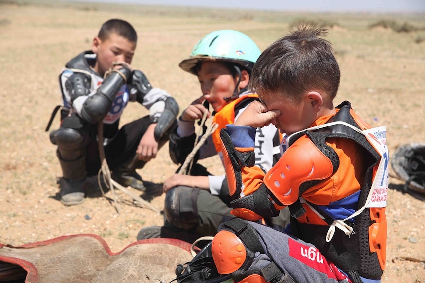 Three child jockeys sits on the ground. One holds his forehead and closes his eyes.