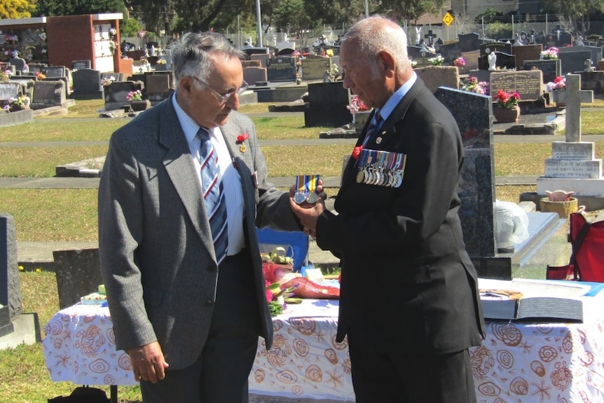 A man wearing medals pinned to his chest hands medals to another man at a cemetery.